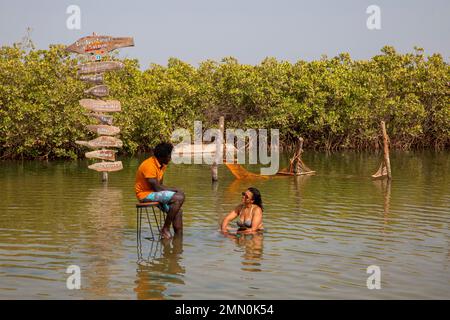 Senegal, Saloum Delta, UNESCO-Weltkulturerbe, Pierre Diouf, Schöpfer der Tourismusagentur Aventuriers du Saloum, sitzt auf einem Hocker neben einer Frau in einer Sandbank im Aventuriers Freizeitcenter du Saloum, das in der Mitte der Mangroven installiert ist Stockfoto