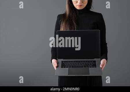 Zugeschnittene Ansicht einer jungen Frau in schwarzem Rollkragenpullover mit Laptop mit leerem Bildschirm isoliert auf dunkelgrau Stockfoto