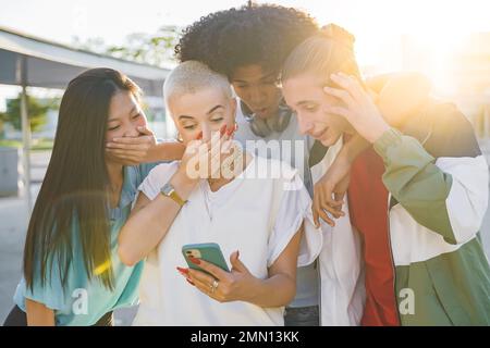 Fröhliche Gruppe von Freunden, die auf ihr Smartphone schauen, überrascht und aufgeregt draußen. Lächelnde junge Leute. Stockfoto