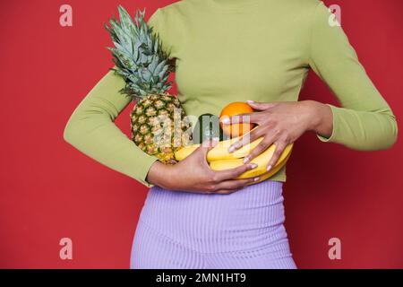 Junge schwarze Frau in grünem Pullover mit isolierten Früchten auf rotem Hintergrund Stockfoto