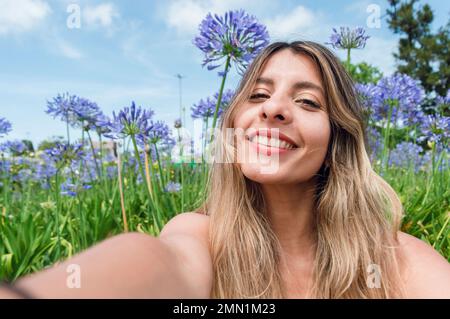 Selfie-Porträt einer wunderschönen jungen Latina, einer kolumbianischen Frau mit langen Haaren, die im Park mit Blumen hinter sich sitzt, sie lächelt und sieht gut aus Stockfoto