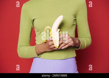 Junge schwarze Frau in grünem Pullover mit isolierter Banane auf rotem Hintergrund Stockfoto