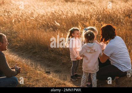 Familie auf einem Spaziergang. Glücklicher Vater sieht seinen dreifarbigen Töchtern draußen beim Spielen mit mom zu Stockfoto
