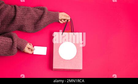 Handgehaltene Kreditkarte und Bastelpaket mit Kopierfeld auf pinkfarbenem Hintergrund. Flach liegen. Geschenke für die Feiertage Stockfoto