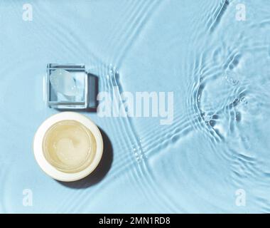 Ein Stück Creme auf Glaspodium mit einem Glas auf Wasserhintergrund. Konzept der feuchtigkeitsspendenden Kosmetika Stockfoto