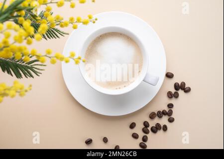 Ein Frühlingsbouquet mit gelben Mimosablüten über einer Tasse Kaffee-Cappuccino auf einem beigen Tisch mit Kaffeebohnen. Konzept vom 8. März, fröhlicher Frauentag Stockfoto