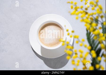 Ein Frühlingsstrauß mit gelben Mimosablüten über einer Tasse Kaffee-Cappuccino auf hellem Hintergrund mit Schatten. Konzept vom 8. März, fröhlicher Frauentag. Stockfoto