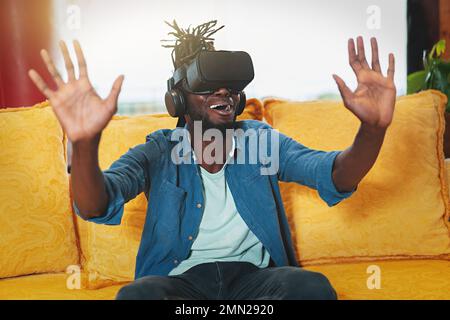 Junger afroamerikanischer Mann, der auf der Couch sitzt, VR-Erfahrung genießt und den Metaverse erforscht. Perfektes Bestandsbild für Technik und Gaming-Konzept Stockfoto