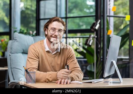 Porträt eines erfolgreichen lächelnden Programmiers im modernen grünen Loft-Büro, eines blonden Mannes, der lächelt und in die Kamera schaut, eines Geschäftsmanns im Pullover und eines legeren Hemds, der in der Nähe mit dem Computer arbeitet. Stockfoto