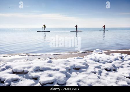 Der Three-man-Athlet paddelt auf dem Sup-Board im See mit Eis im Winter und Schnee am Strand. Extremes Outdoor-Sportkonzept Stockfoto