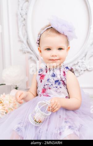 Elegantes Kleid für ein kleines Mädchen. Ein einjähriges Mädchen in einem geschwollenen Kleid und einer süßen Schleife posiert vor dem Hintergrund eines hellen Raumes mit Frisiertisch und Stockfoto