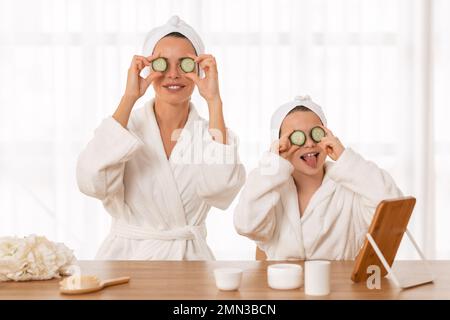 Fröhliche Mutter und kleine Tochter, die Bademäntel tragen und Gurkenscheiben auf die Augen kleben Stockfoto