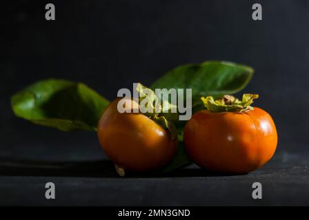 Reife orangefarbene Persimonen, verzehrfertig auf dunklem Hintergrund und horizontal beleuchtet Stockfoto