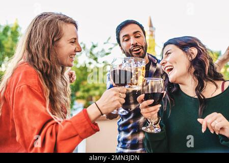 Zwei junge Frauen und ein junger Mann haben eine tolle Zeit beim Witzen und Toasten mit Wein- und Bierbechern im Freien Stockfoto