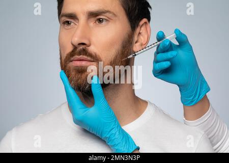 Ein bärtiger Mann bekommt eine Schönheitsinjektion, ein Kosmetiker mit Handschuhen, der Anti-Aging-Verfahren für den Menschen durchführt Stockfoto