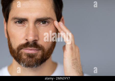 Gesichtspflege bei Männern. Bärtiger Mann mittleren Alters, der das Gesicht berührt, Haut unter den Augen pflegt, grauer Hintergrund, Kopierraum Stockfoto