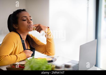 Glückliche afroamerikanische Millennialfrau in der Schürze, die Fischgericht probiert, Finger leckt, sich einen Video-Blog auf dem Laptop ansieht Stockfoto