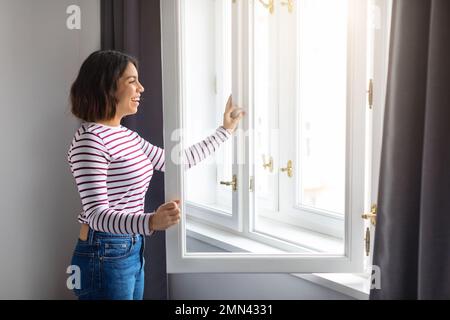 Porträt Einer Lächelnden Arabischen Jungen Frau, Die Zu Hause Das Fenster Öffnet Stockfoto