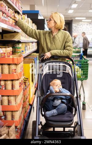 Casualy kleidete die Mutter, die im Supermarkt Konserven auswählte, mit ihrem kleinen Jungen im Kinderwagen Stockfoto