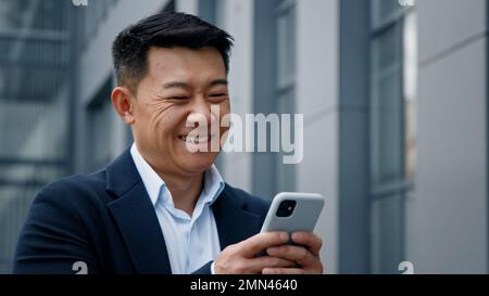 Emotionales Lächeln Asiens im mittleren Alter koreanischer Geschäftsmann Arbeitgeber Unternehmer lacht, wenn er auf ein Mobiltelefon schaut, sich chattet und lächelt Stockfoto