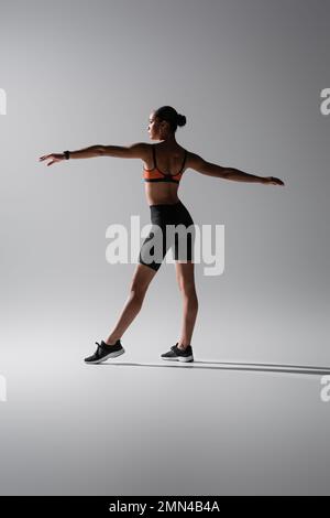 Tätowierte afroamerikanische Frau in Sport-BH und Shorts mit ausgestreckten Händen auf Grau Stockfoto