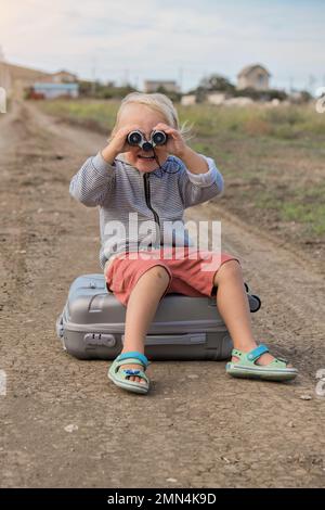 Das Kind schaut durch das Fernglas und sitzt auf einem Koffer vor dem Hintergrund der Straße. Ein Reisender, der im Urlaub spielt. Hochwertiges Foto Stockfoto