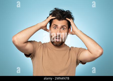 Ein Mann mittleren Alters mit unausgeprägter Frisur, der Haare berührt und schockiert in die Kamera schaut, über blauem Hintergrund steht Stockfoto