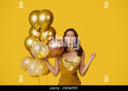 Konzept der Feiertagsfeier. Eine glückliche Frau in einem Kleid, die einen Haufen Ballons hält und sich was wünscht, und auf gelbem Hintergrund posiert Stockfoto