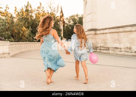 Tochter Mutter lief und hielt Händchen. In blauen Kleidern mit fließenden langen Haaren vor dem Hintergrund eines Sonnenuntergangs und einem weißen Gebäude. Stockfoto