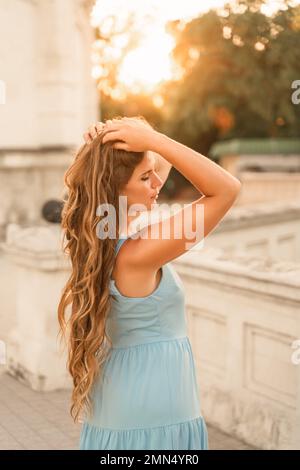 Ein blaues Kleid für Frauen. Porträt einer Frau mit langen Haaren und einem blauen Kleid vor dem Hintergrund der untergehenden Sonne und eines weißen Gebäudes. Lifestyle Stockfoto