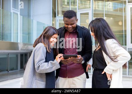 Attraktive Kollegen, die ein Tablet außerhalb des Büros verwenden. Gruppe junger Freunde, die bei der Arbeit in der Stadt lächeln. Ethnische Vielfalt, soziale Eingliederung Stockfoto