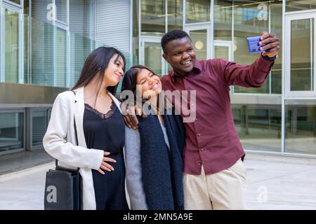 Attraktive Kollegen machen ein Selfie mit einem Smartphone außerhalb des Büros. Gruppe von Freunden, die in der Arbeitspause lächeln. Ethnische Vielfalt, soziale Eingliederung. Stockfoto