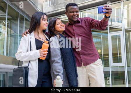 Attraktive Kollegen machen ein Selfie mit einem Smartphone außerhalb des Büros. Gruppe von Freunden, die in der Arbeitspause lächeln. Ethnische Vielfalt, soziale Eingliederung. Stockfoto