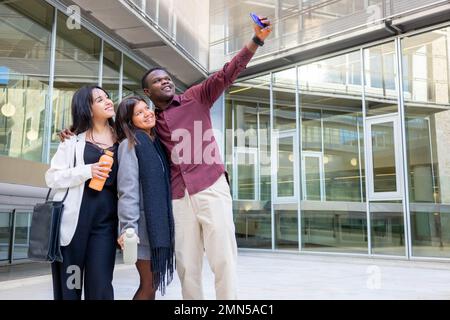 Attraktive Kollegen machen ein Selfie mit einem Smartphone außerhalb des Büros. Gruppe von Freunden, die in der Arbeitspause lächeln. Ethnische Vielfalt, soziale Eingliederung. Stockfoto