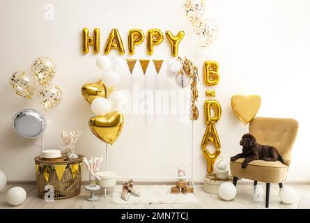 Schokoladen-Labrador-Retriever und Phrase HAPPY BIRTHDAY hergestellt aus goldenen Ballon-Buchstaben im dekorierten Zimmer Stockfoto