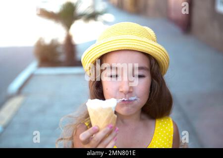 Das Kind isst im Sommer im Freien köstliches Eis und verschmutzt seinen Mund. Ein Mädchen mit gelbem Hut und ein Sonnenkleid in der Hitze eines clos Stockfoto