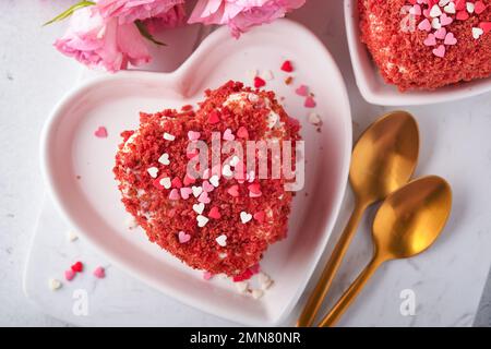 Zwei Kuchen Roter Samt in Form von Herzen auf weißem Teller, Rosenblumen und eine Tasse Kaffee auf rosafarbenem romantischem Hintergrund. Dessert-Idee für den Valentinstag, Stockfoto