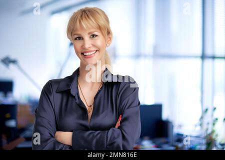 Glückliche Geschäftsfrau mit blonden Haaren, die im Büro mit gefalteten Armen stand. Stockfoto