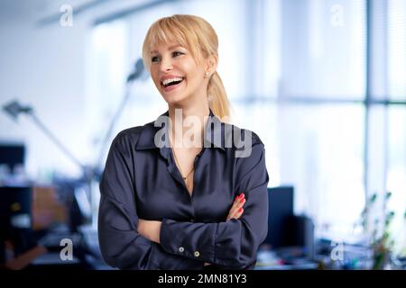 Glückliche Geschäftsfrau mit blonden Haaren, die im Büro mit gefalteten Armen stand. Stockfoto