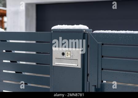 Ein modernes Videotelefon mit einem Briefkasten und einem drahtlosen Kartenleser, montiert in einem Anthrazit-Zaun, sichtbares Wicket, es regnet im Winter. Stockfoto