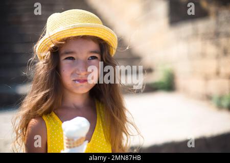 Das Kind isst im Sommer im Freien köstliches Eis und verschmutzt seinen Mund. Ein Mädchen mit gelbem Hut und ein Sonnenkleid in der Hitze eines clos Stockfoto