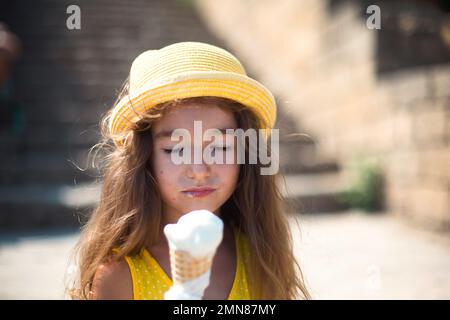 Das Kind isst im Sommer im Freien köstliches Eis und verschmutzt seinen Mund. Ein Mädchen mit gelbem Hut und ein Sonnenkleid in der Hitze eines clos Stockfoto