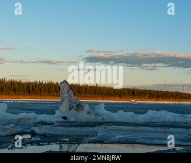 Eine Eisscholle in Form einer Haifischflosse ragt vor dem Hintergrund von Stromleitungen und Wäldern aus der Oberfläche eines schmelzenden Flusses heraus. Stockfoto