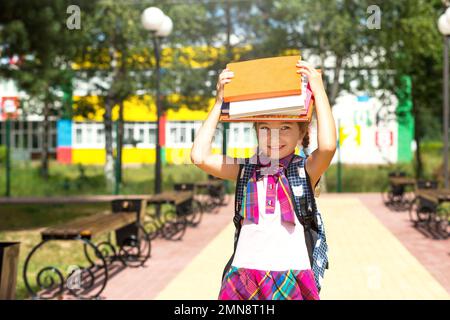 Mädchen mit einem Rucksack und einem Stapel Bücher auf dem Kopf in der Nähe der Schule. Zurück in die Schule, das Kind ist müde, schwere Lehrbücher. Bildung, Grundschule Cla Stockfoto