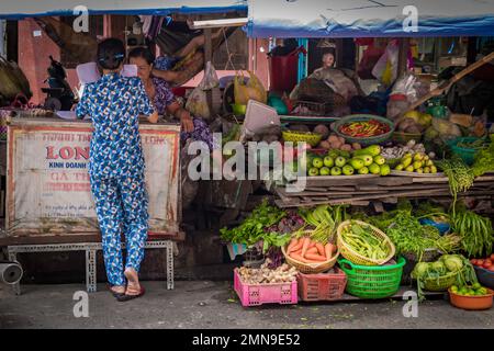 Rücken einer Verkäuferin, die vor einem Stall mit Obst und Gemüse in Vietnam steht. Stockfoto