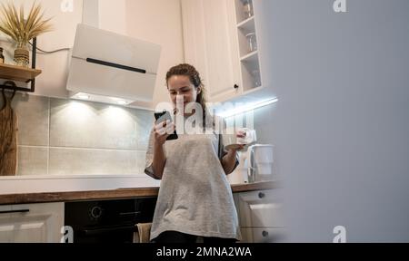 Junge Frau, die in der Küche steht, eine Kaffeetasse hält und ihr Handy benutzt. Stockfoto
