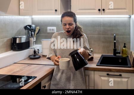 Eine Lifestyle-Frau am Morgen steht in der Küche, gießt Kaffee in eine Tasse aus der türkischen Kaffeemaschine und gähnt. Stockfoto