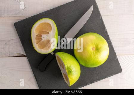 Zwei Hälften und ein ganzer natürlicher Bio-Citrus Sweetie auf einem Schiefertafel mit einem Metallmesser, Nahaufnahme, auf einem weißen Holztisch. Stockfoto