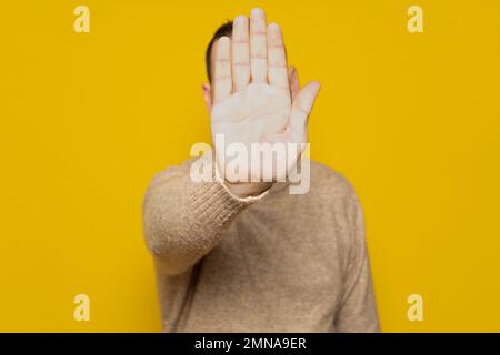 Hispanischer Mann, der die Hand nach vorne legt, um die Kamera anzuhalten, und Fotos oder Bilder gegen die gelbe Wand ablehnt. Stockfoto