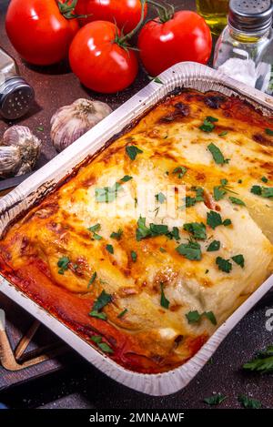 Hausgemachte Cannelloni-Pasta, großes Tablett mit gebackenen Cannelloni mit Hackfleisch mit Béchamel-Sauce, mit Kräutern in Zutaten auf dunklem Hintergrund Stockfoto